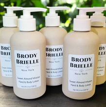 Load image into Gallery viewer, Row of Brody Brielle hand and body lotion bottles on a wooden surface with a blurred green background.
